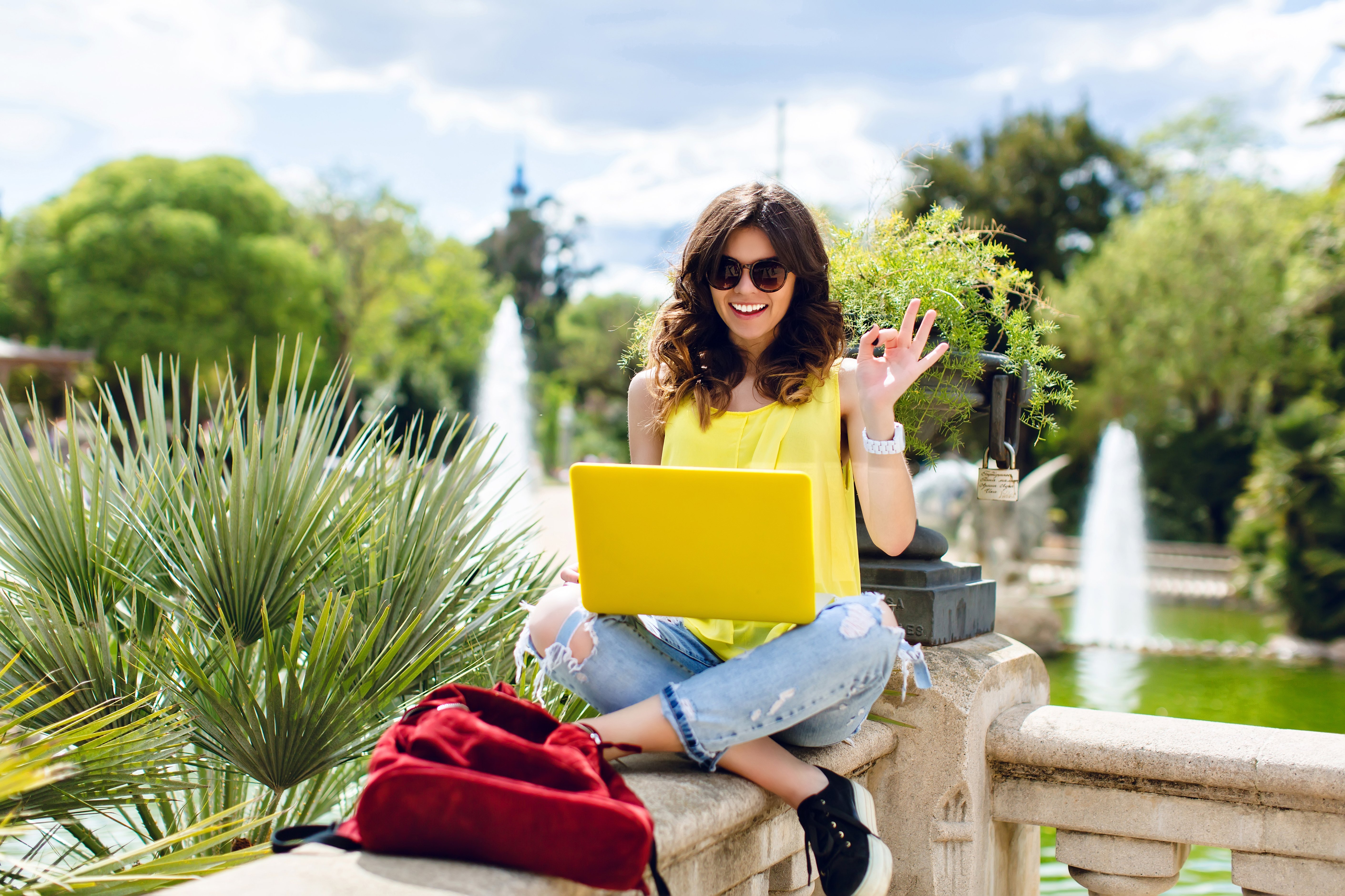 brunette-girl-sunglasses-is-sitting-fence-park-she-holds-yellow-laptop-smiling-camera