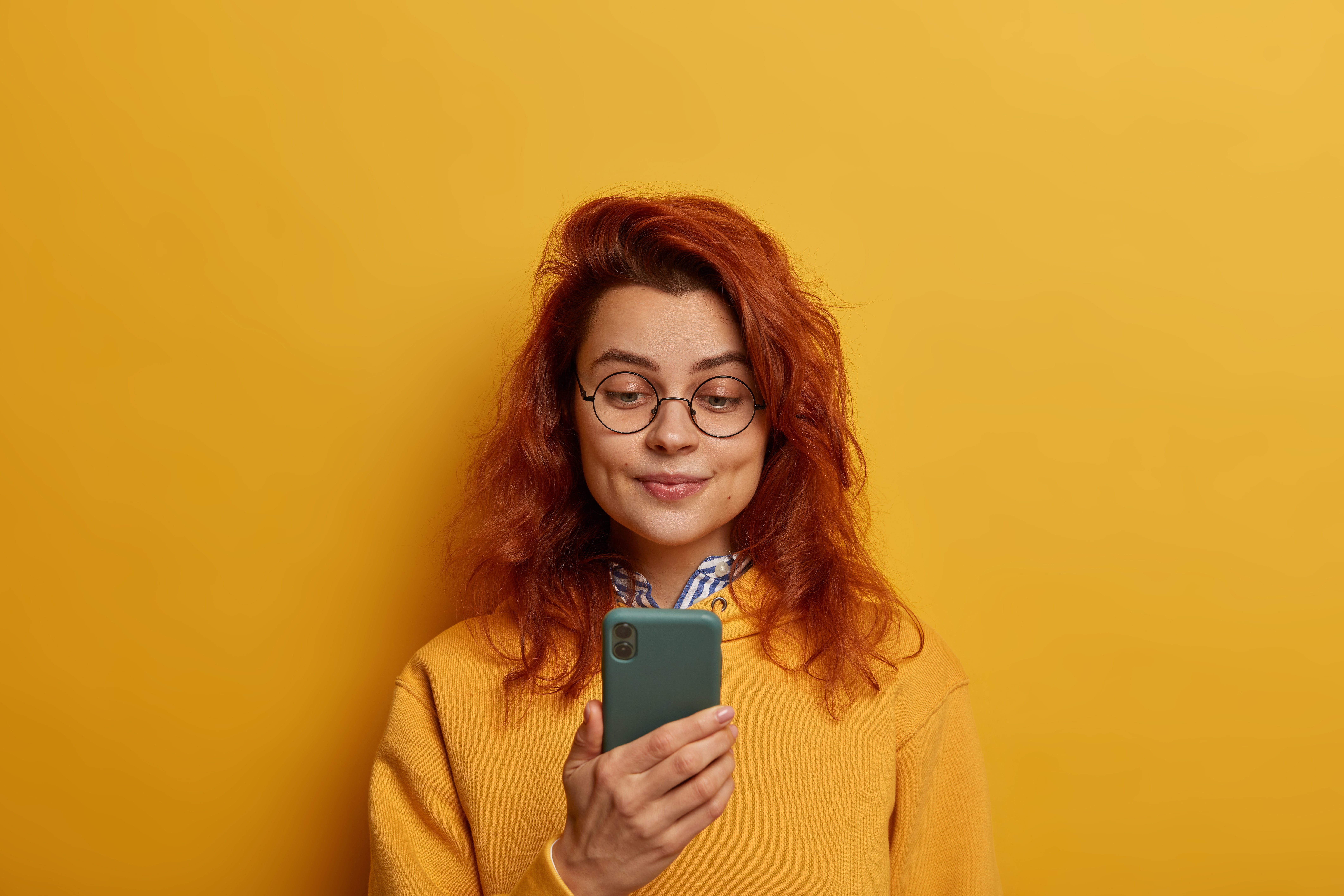 ginger-young-woman-holds-mobile-phone-reads-notification-wears-round-glasses-yellow-jumper
